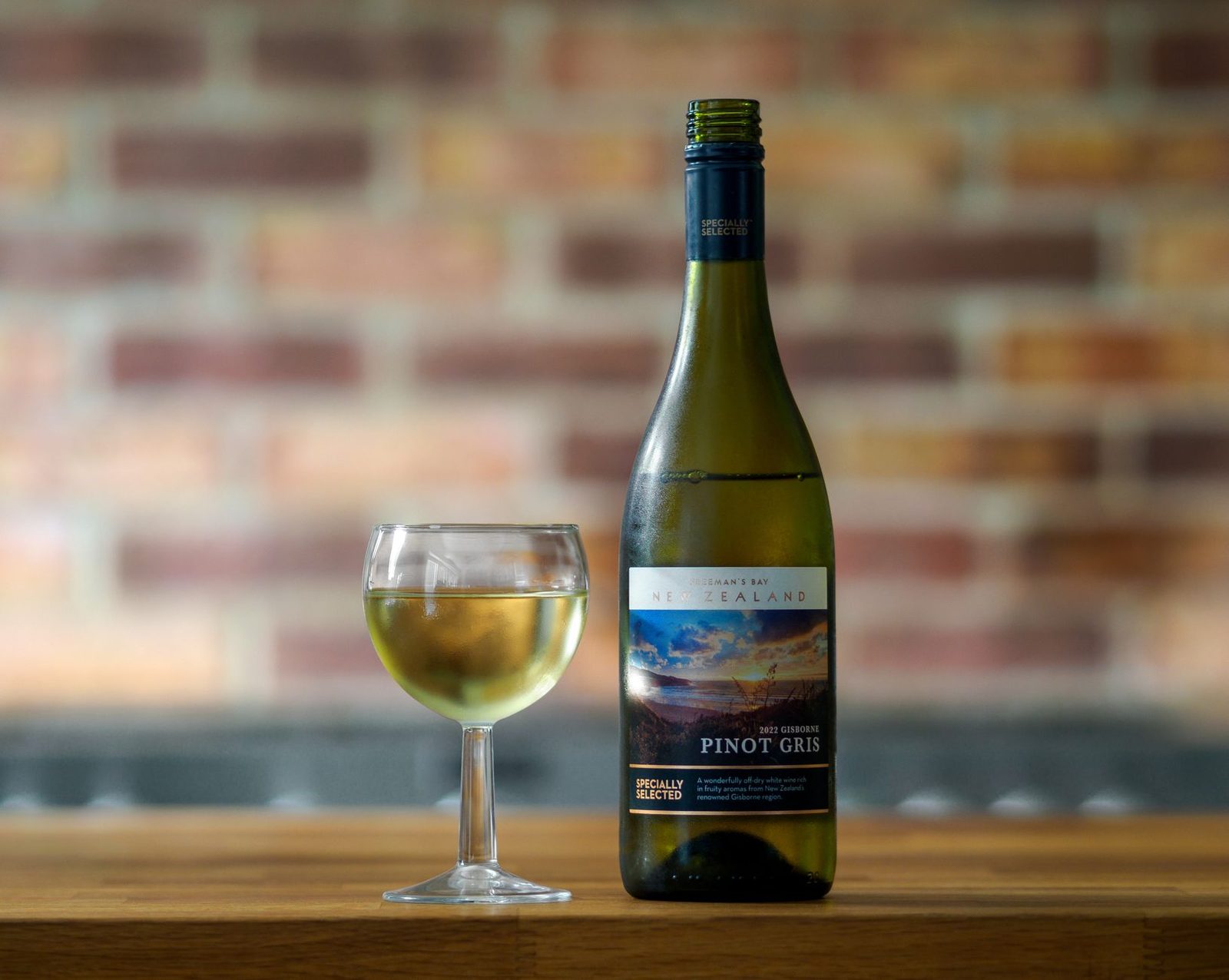 Close-up of a New Zealand Pinot Gris wine bottle and glass set against a blurred brick wall.