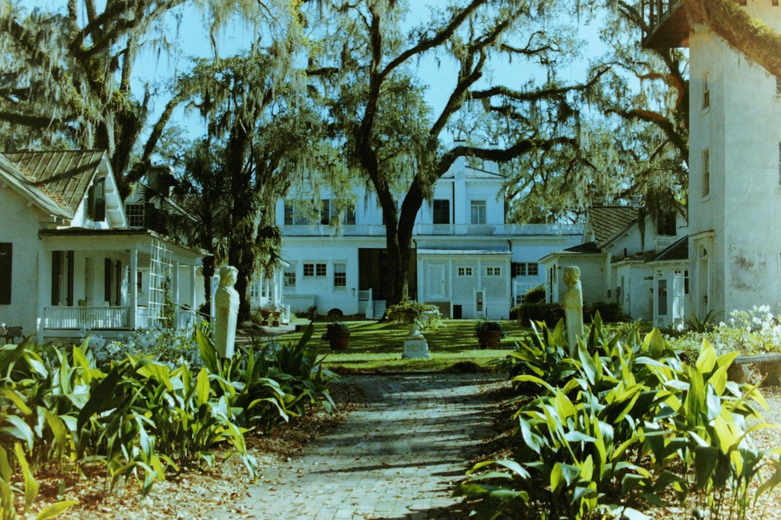 A serene view of a historic manor surrounded by a lush garden with statues and Spanish moss.