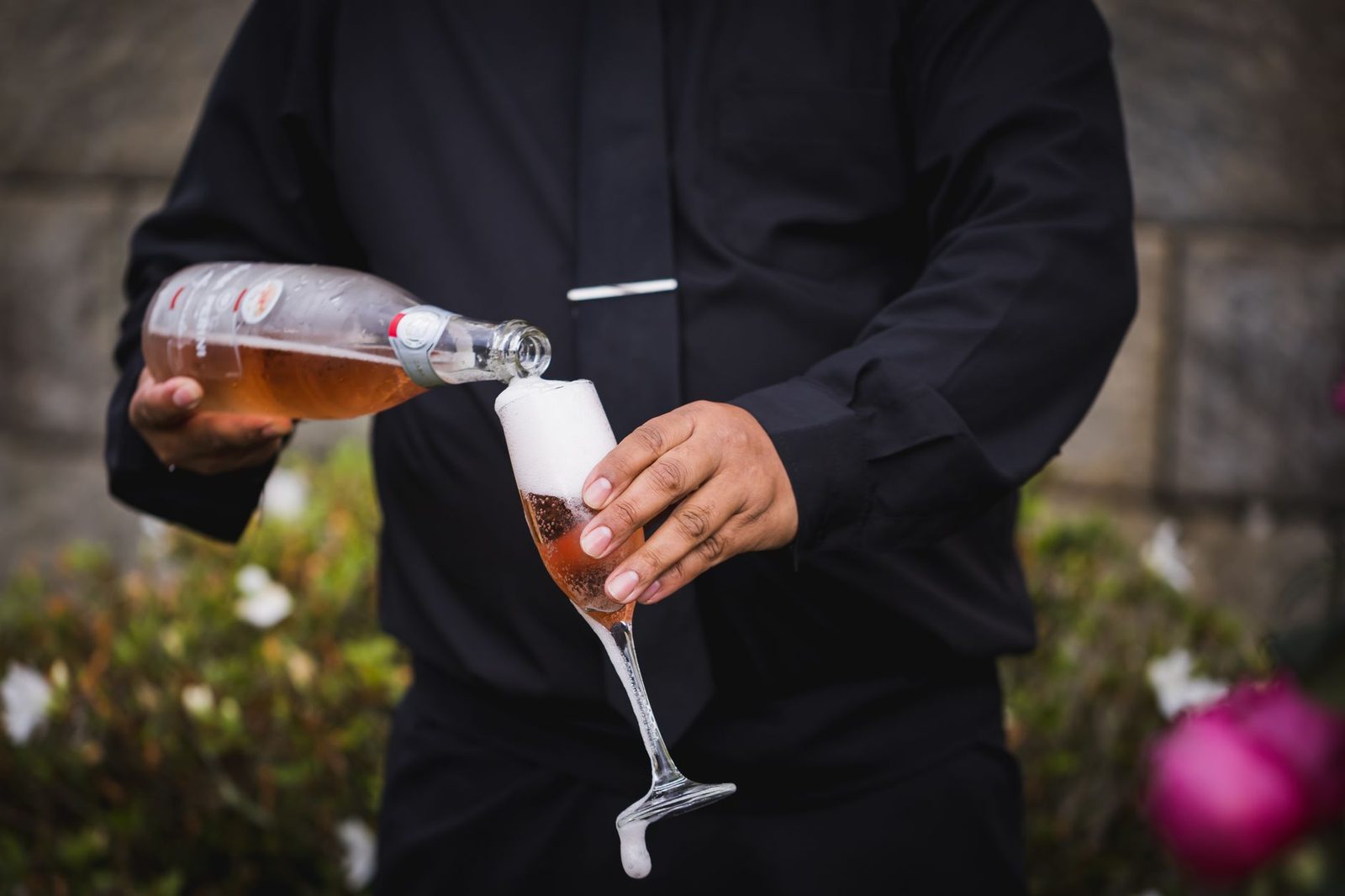 Close-up of rosé champagne being poured into a flute by a person in formal attire outdoors.