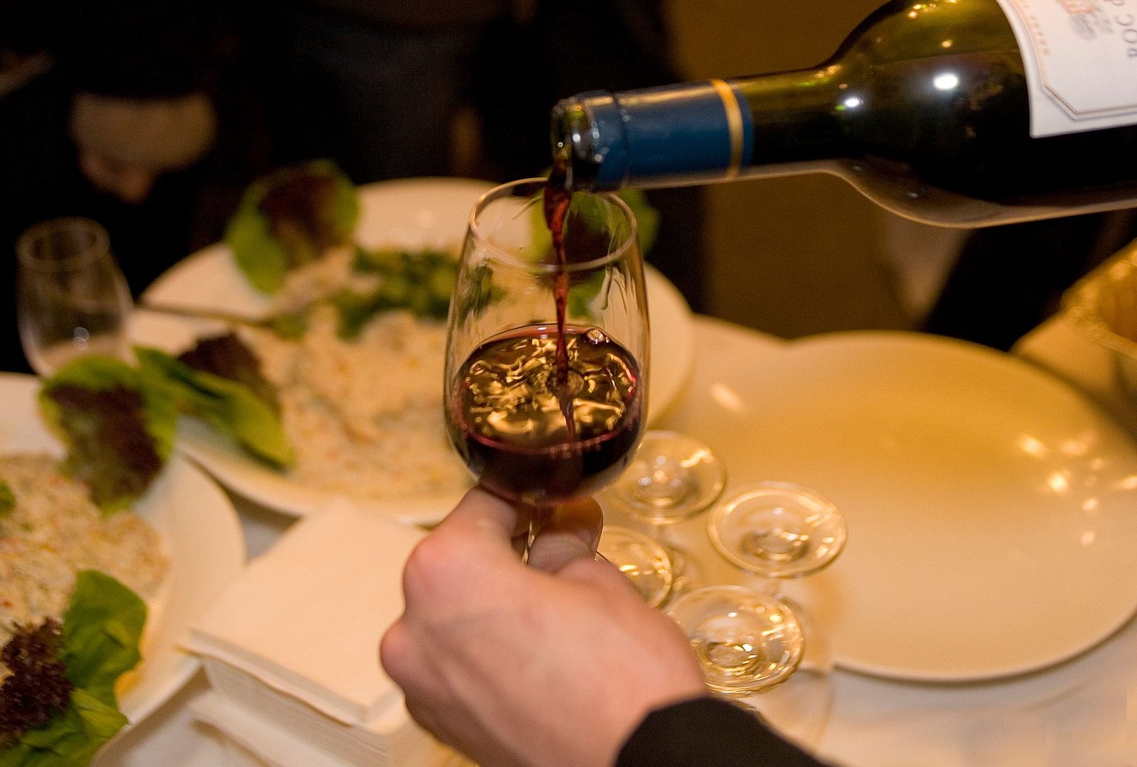 Close-up of red wine being poured into a glass at an elegant dinner with lush salads on the table.