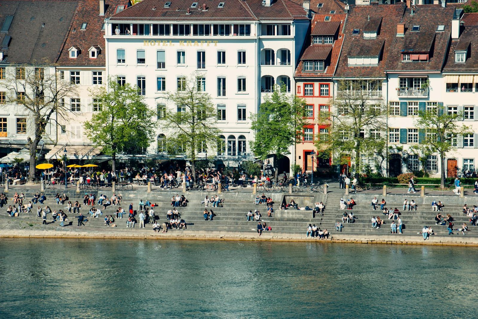 People relax and socialize by the Rhine River in Basel, showcasing vibrant city life.