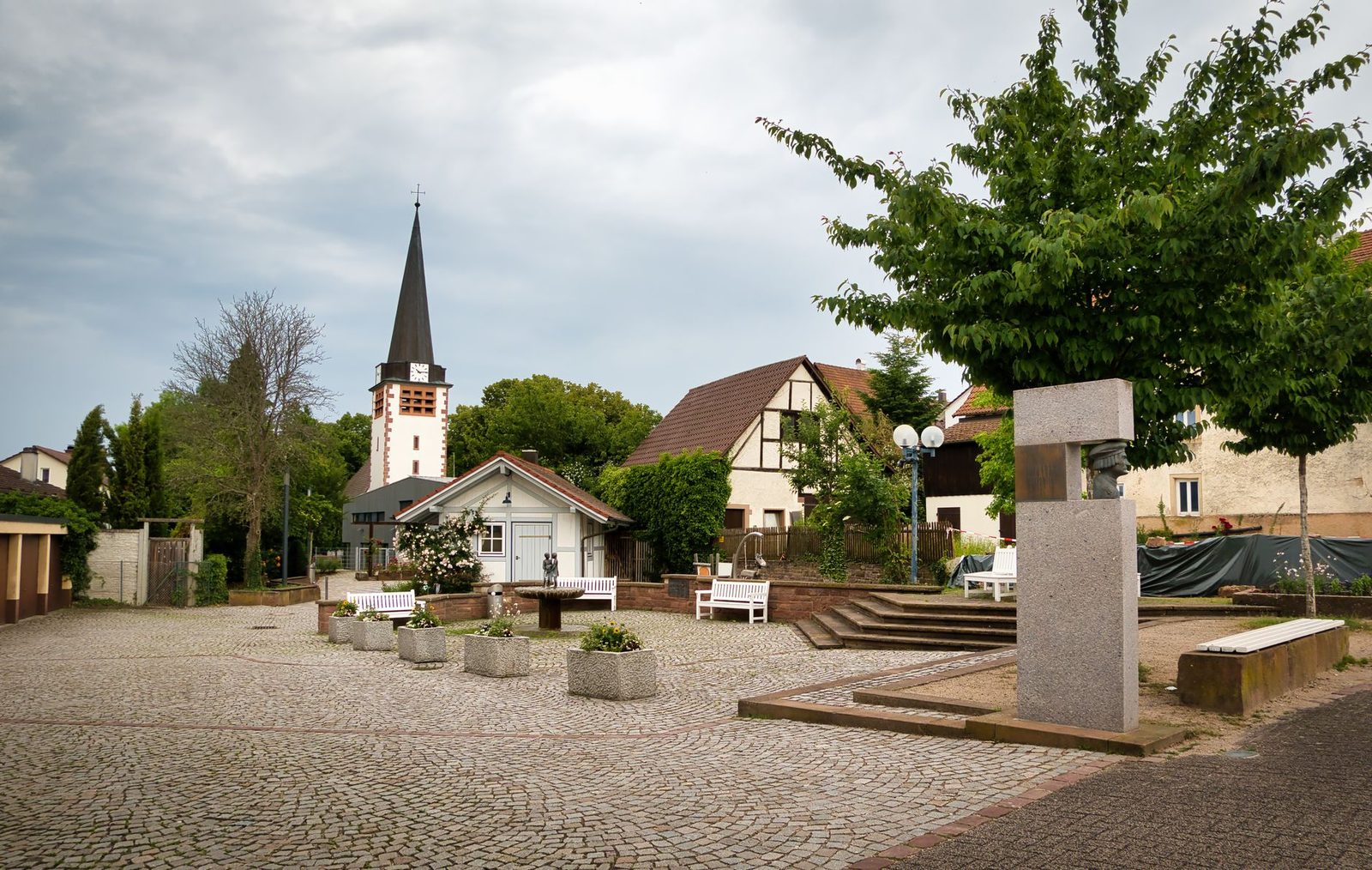 Quaint village square in Pforzheim with church and benches under a cloudy sky.
