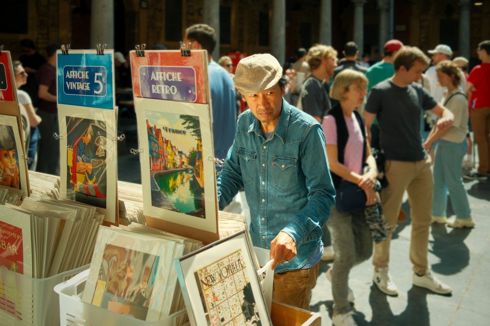 A man in denim casually examines vintage posters at a bustling outdoor market in Lille, France.