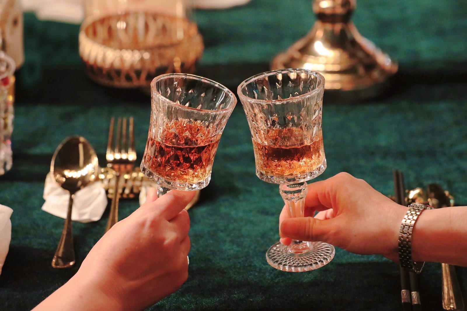 Two hands toasting with crystal glasses filled with amber liquid at a formal dinner setting.