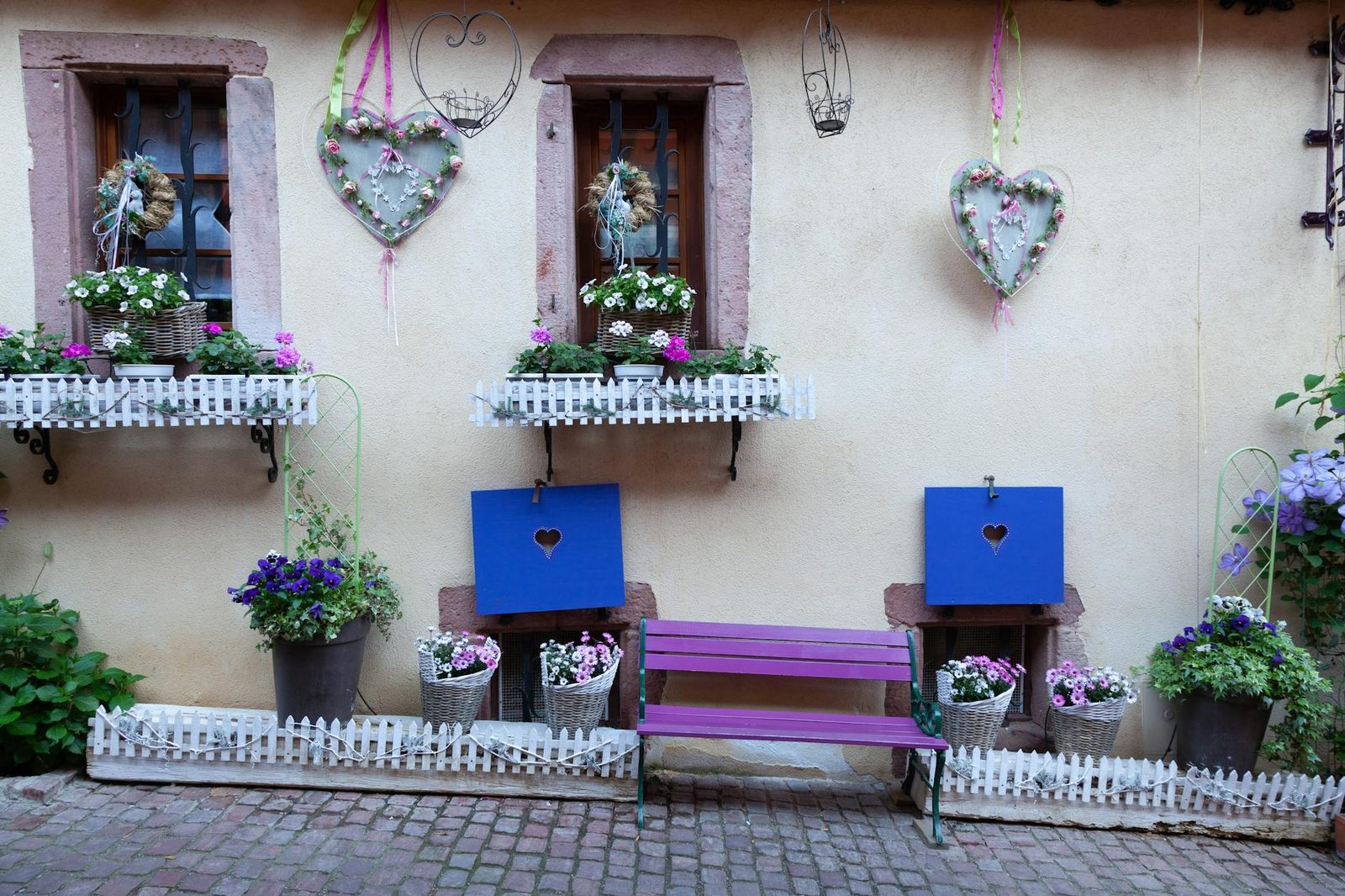 Charming French window scene with flowers, heart decorations, and a purple bench in Riquewihr.