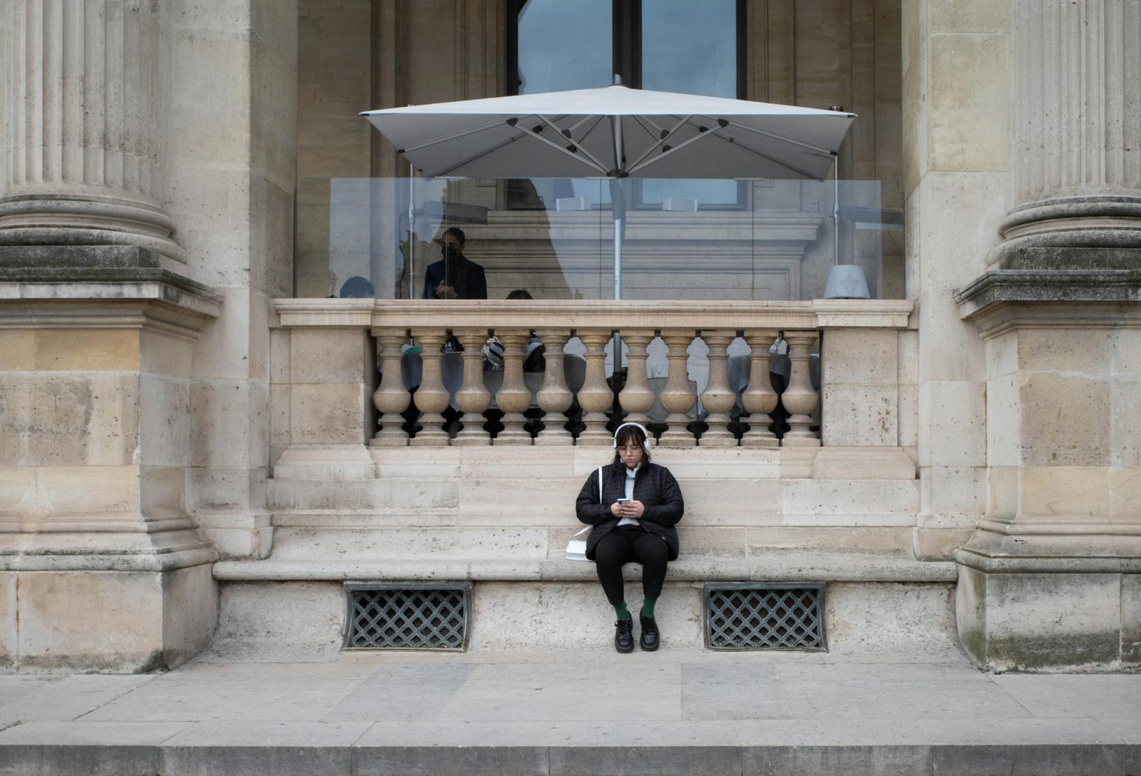 Woman wearing headphones sitting on stone steps in front of historic building, city setting.