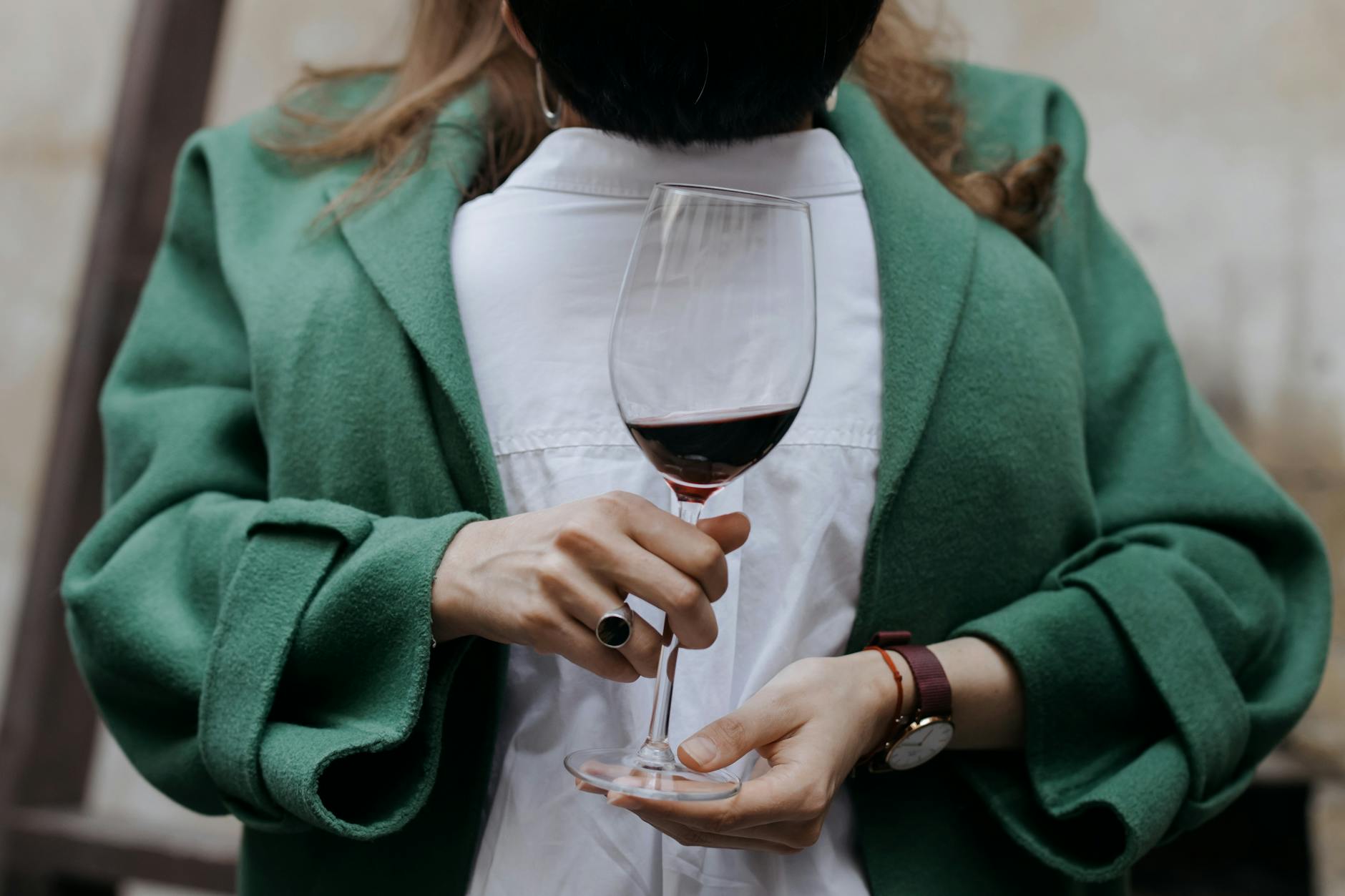 A woman in a green coat holds a wine glass filled with red wine, showcasing elegance and style.