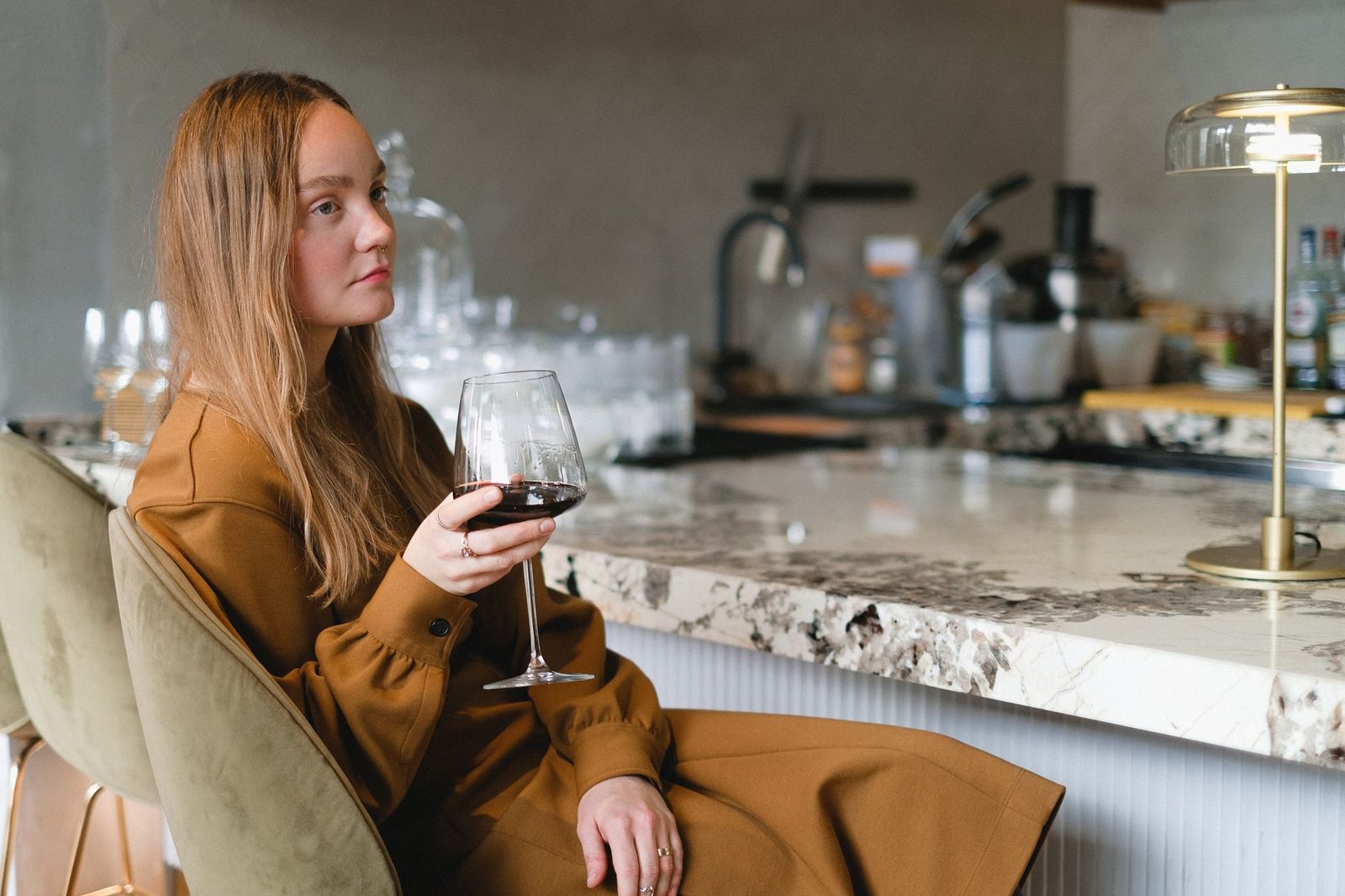 A contemplative woman in a brown dress holding a wine glass at a marble counter.